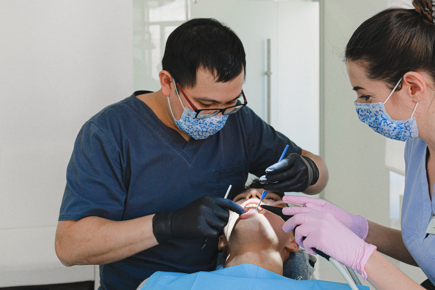 A dentist and an assistant wearing masks and gloves perform a dental procedure on a patient in a clinical setting.