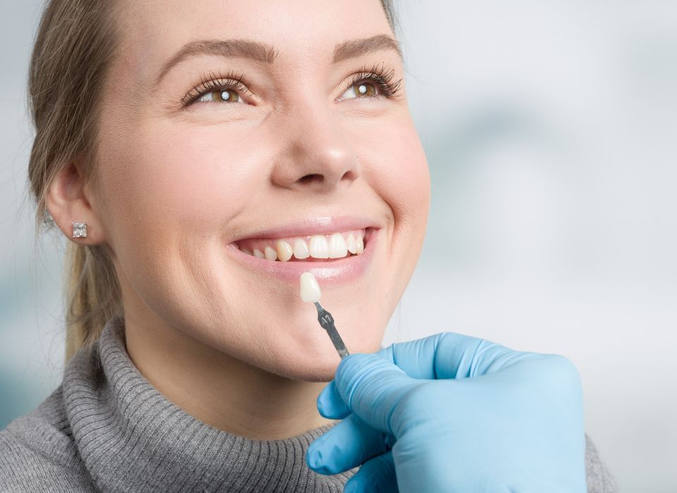 A dental professional in blue gloves holds a tooth shade guide against a patient's teeth to match their color.