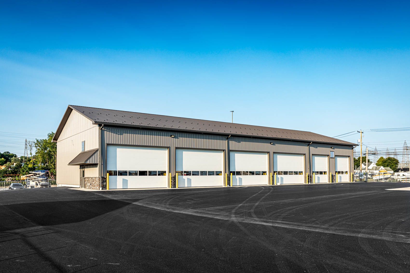 Tan metal building with multiple white garage doors, asphalt parking area, blue sky.
