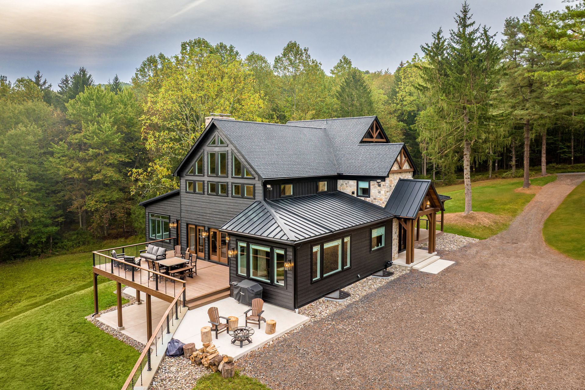 Cabin with dark siding, surrounded by trees, with deck and gravel driveway.