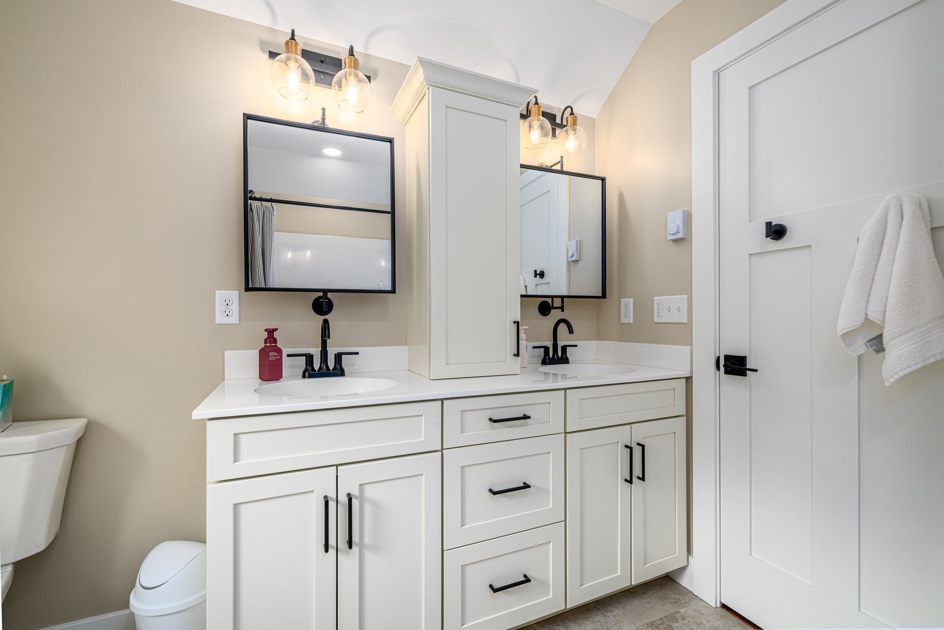 White bathroom with double vanity, mirrors, and door; neutral walls, black fixtures.