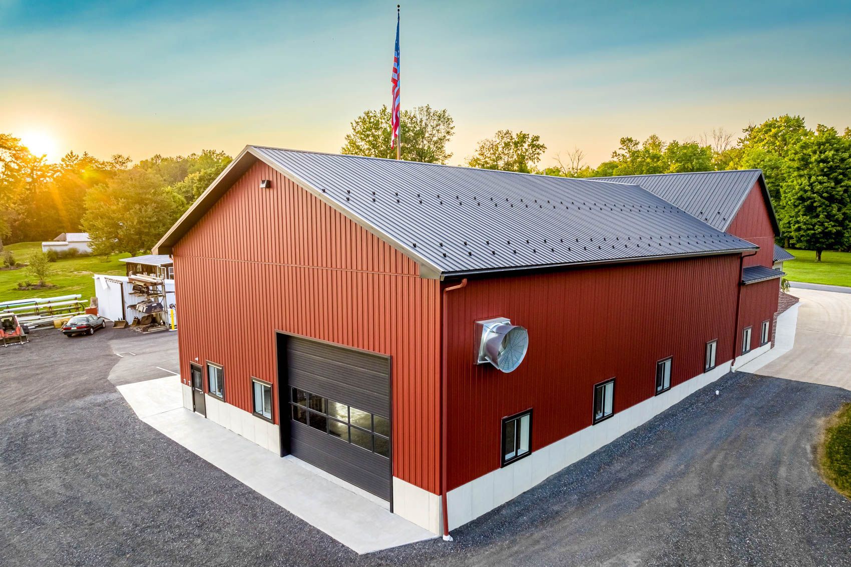 Red barn-style building with a gray roof and large garage door. A flagpole and setting sun are in the background.