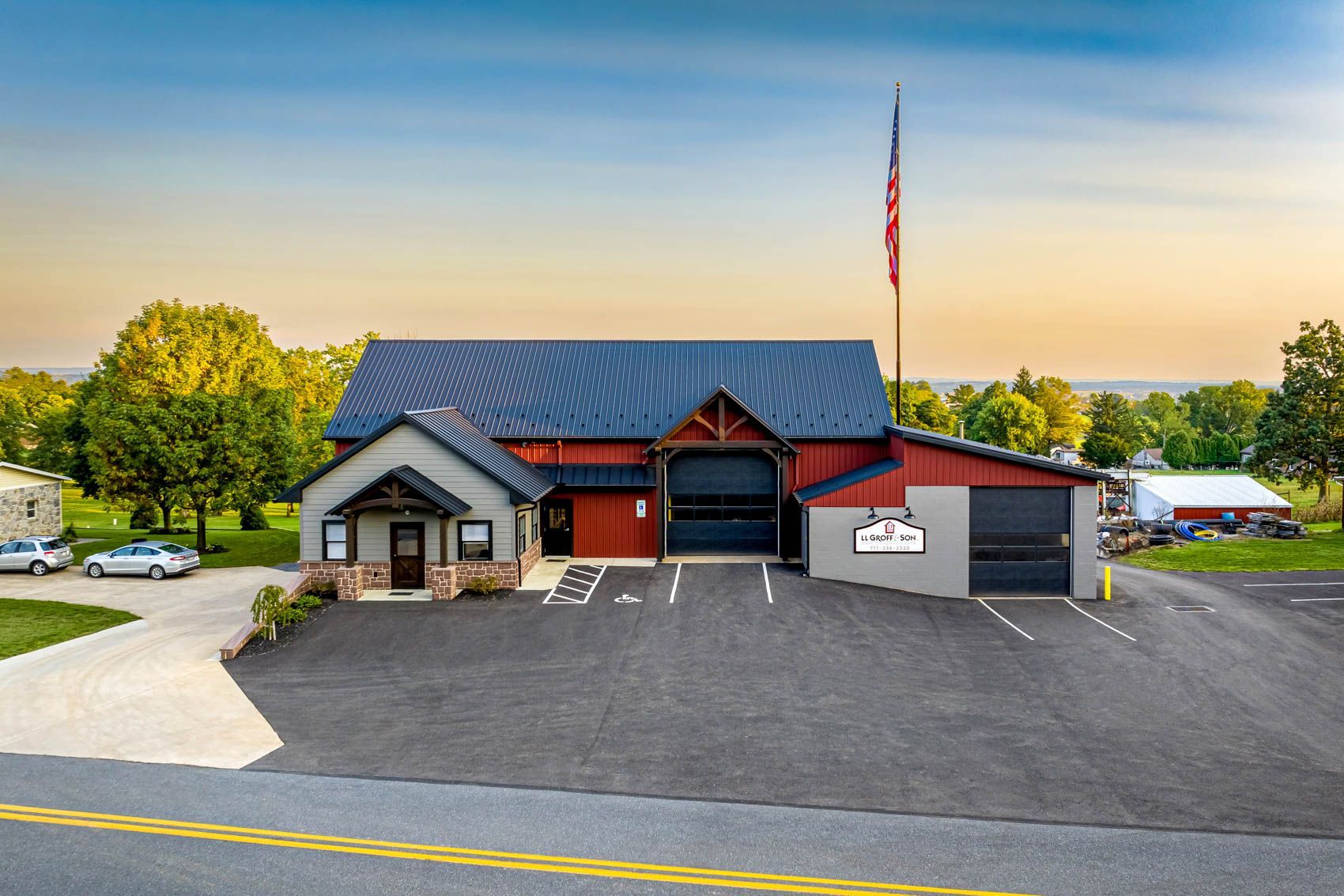Fire station with American flag, dark roof, red and gray exterior, parked cars, and trees.