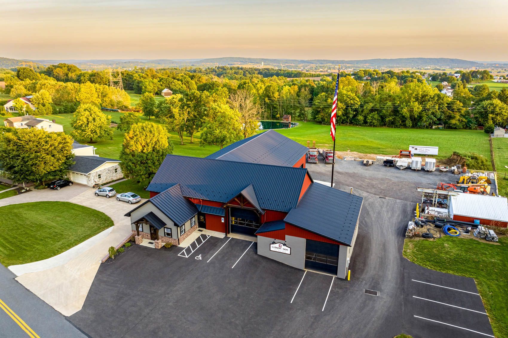 Fire station with red and gray exterior, parked vehicles, American flag, and surrounding rural landscape.