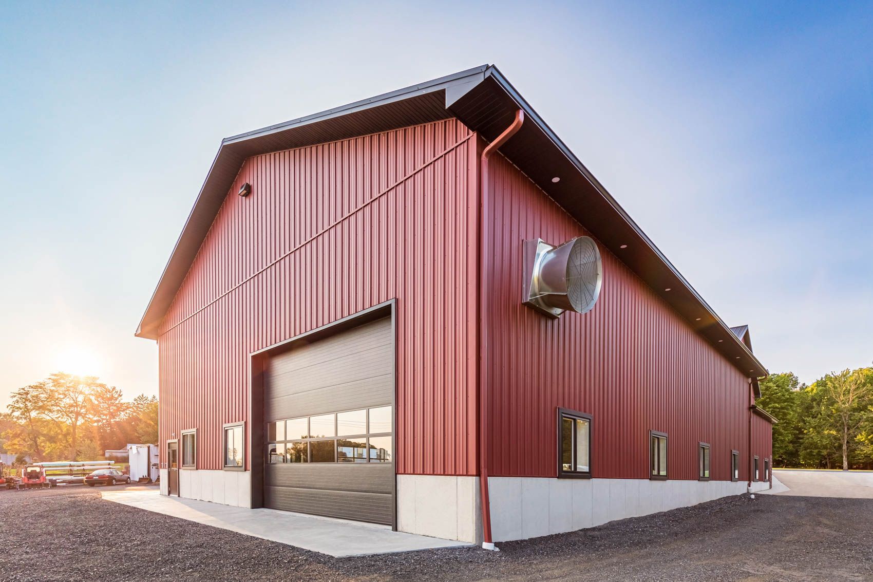Red metal barn with large garage door, set on a gravel base under a blue sky.