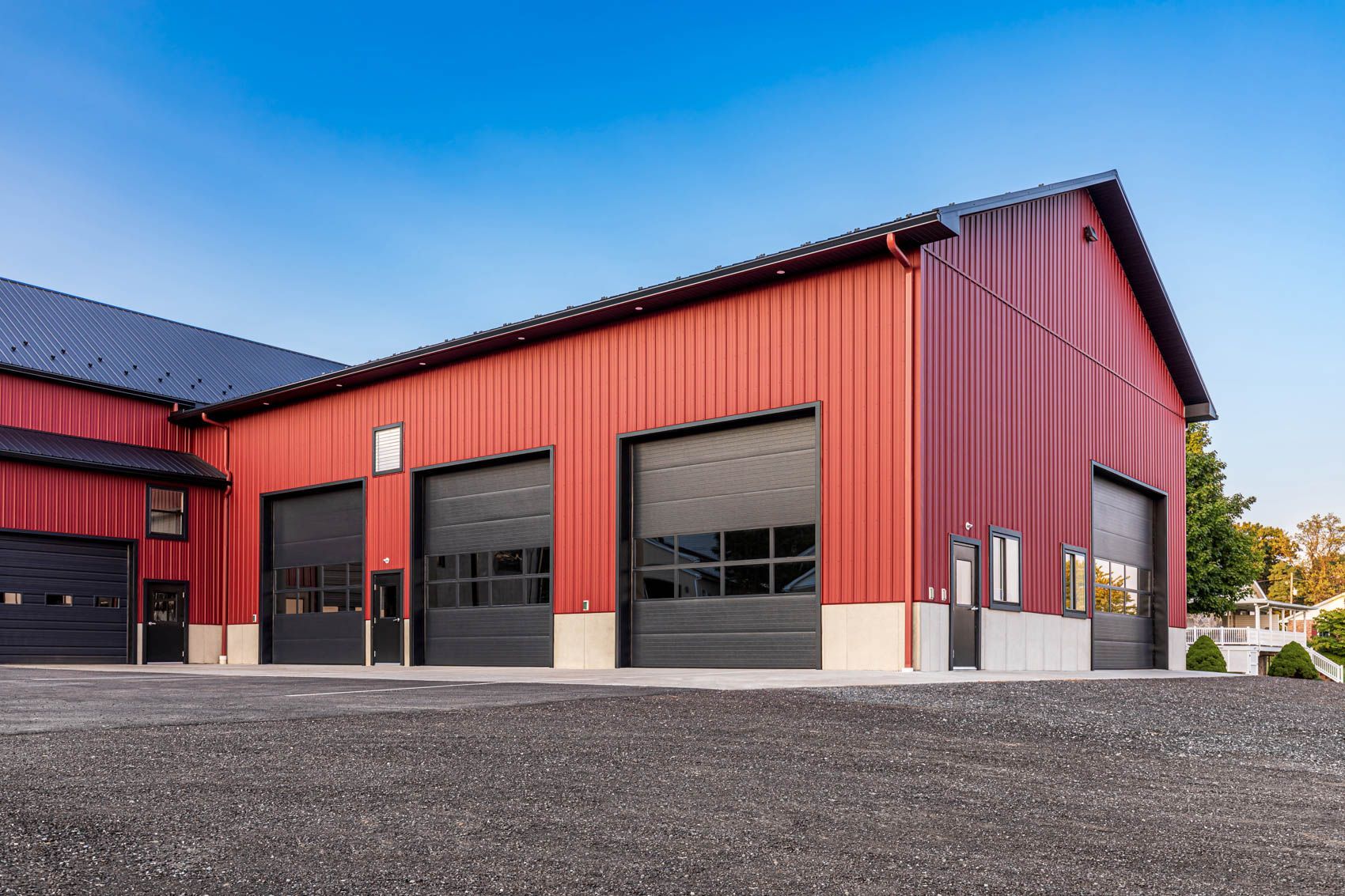 Red metal building with multiple black garage doors and a gravel driveway under a blue sky.