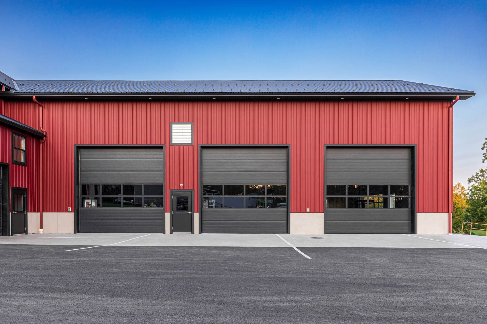 Red building with three garage doors and small door, black roofs, and gravel surface.