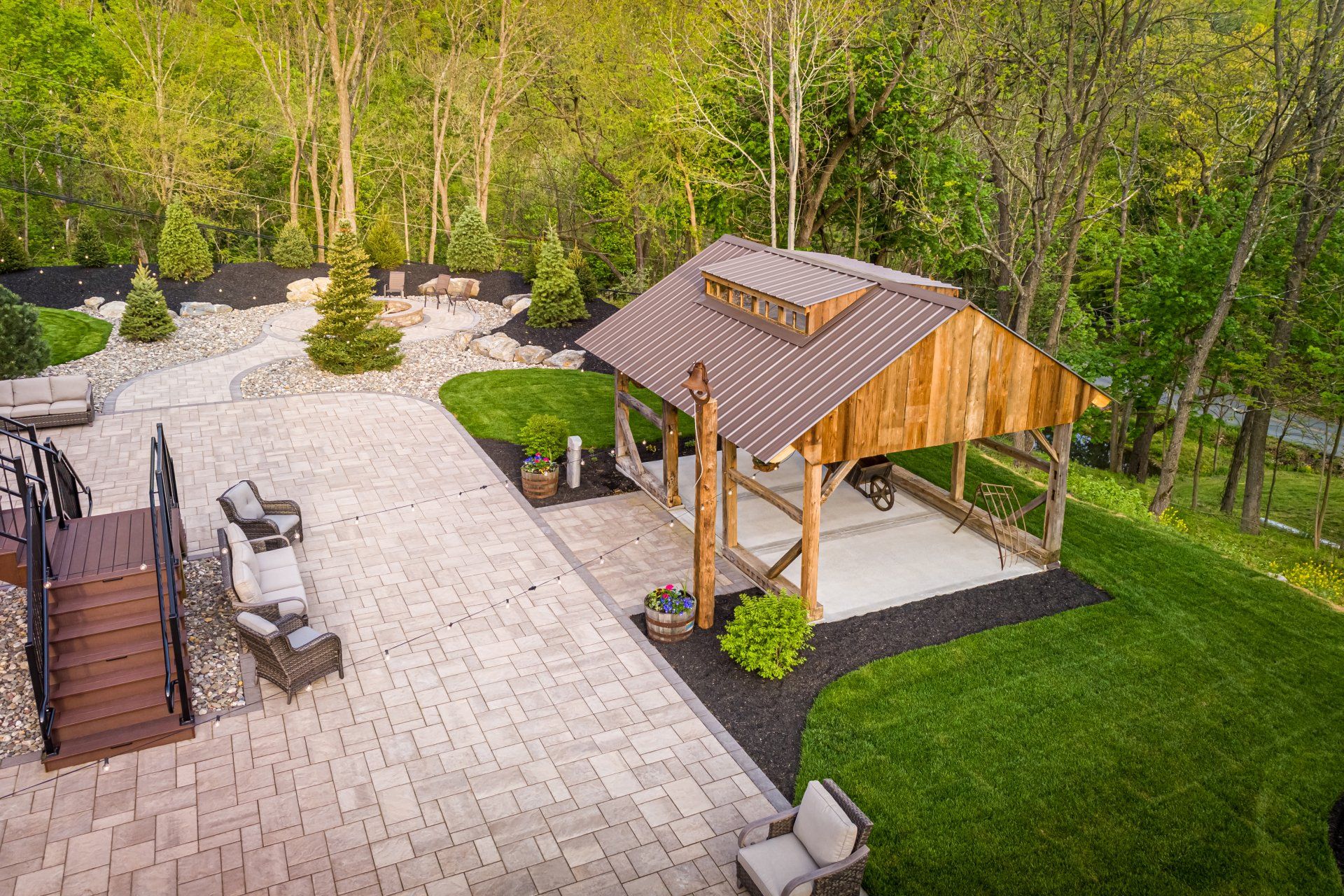 Patio with wooden shelter, seating, pavers, and green lawn surrounded by trees.