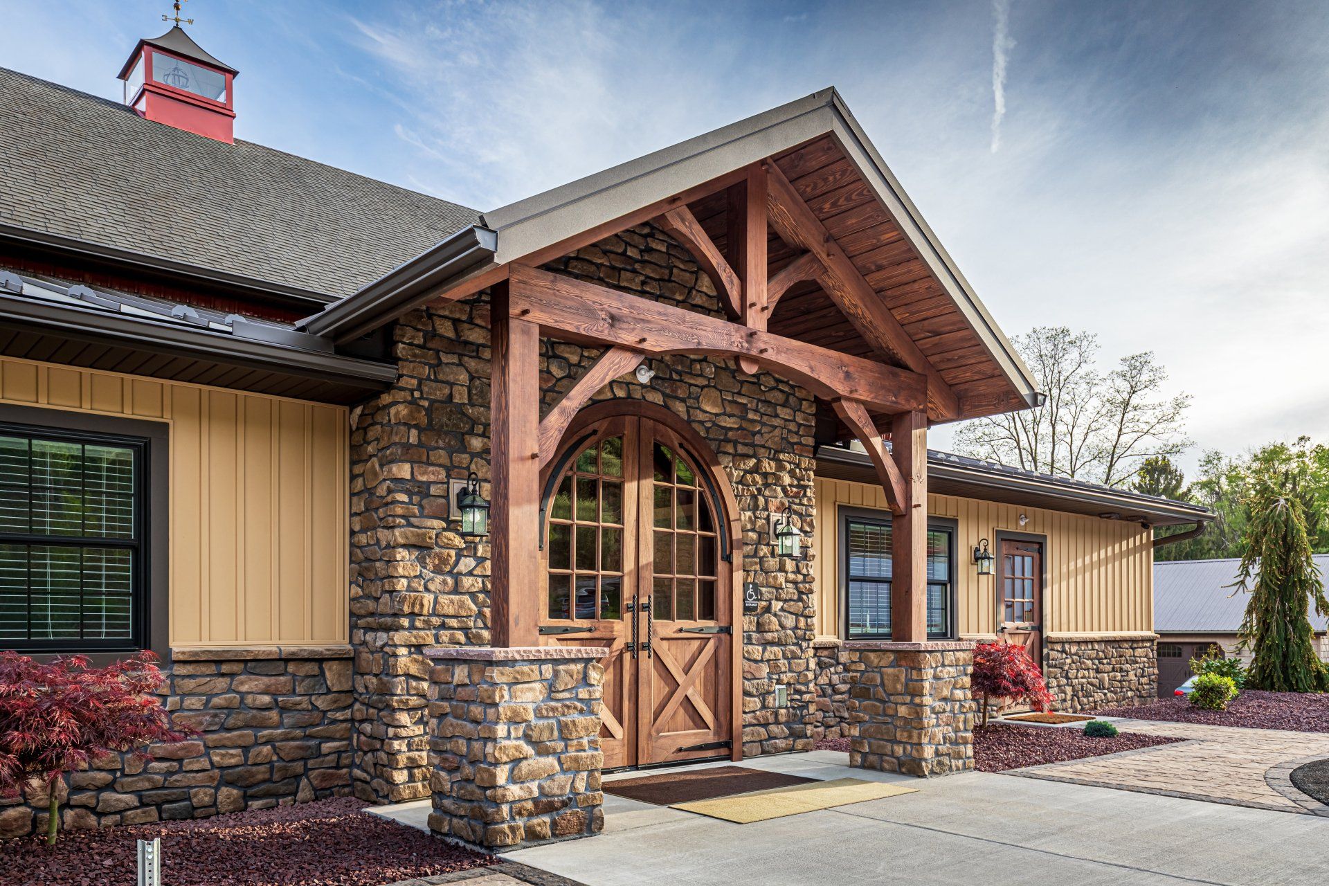 Stone and wood building entrance, arched door, wooden beams, tan siding, red roof.