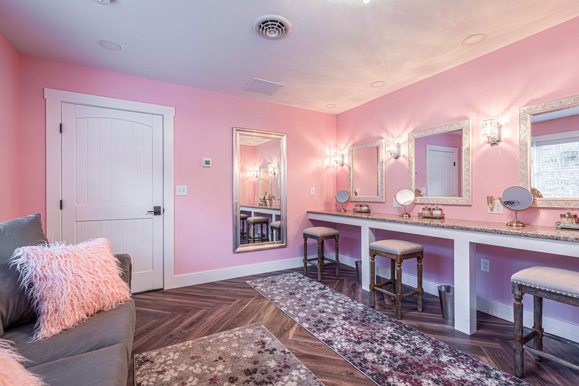 Pink-walled dressing room with vanities, mirrors, seating, and a sofa with pink fluffy pillows.