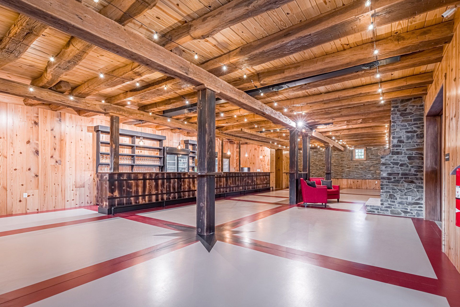 Interior of a rustic event space with wooden beams, red and white floor, bar, and stone wall.