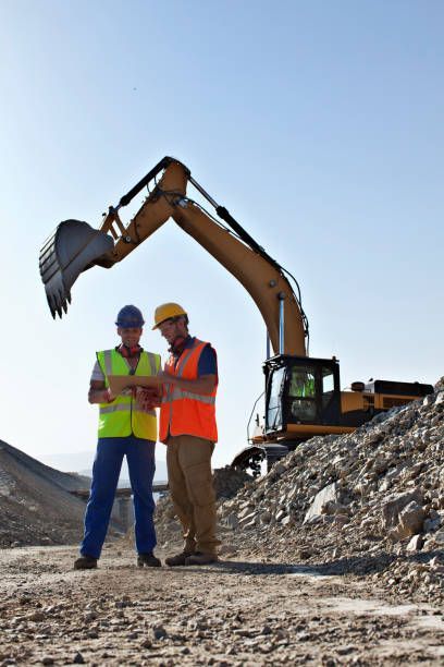 Two construction workers in safety vests review a tablet near an excavator on a worksite.