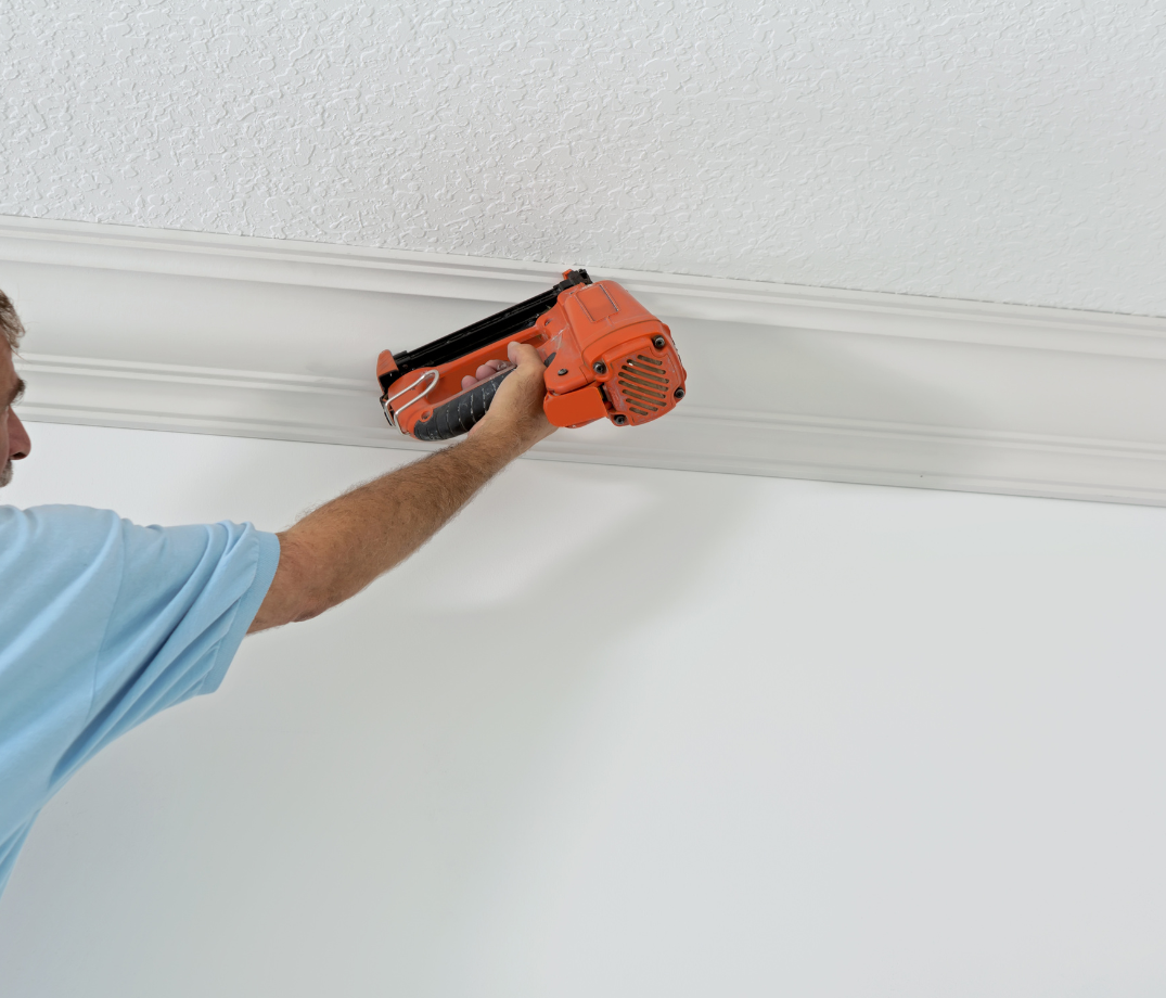 Person using a nail gun to install white crown molding on a white wall.