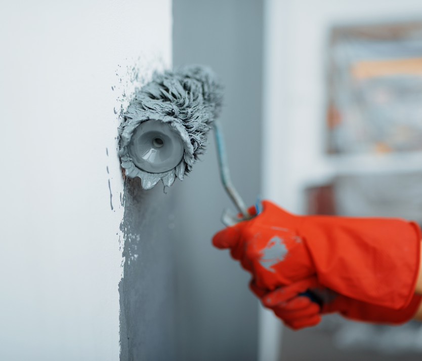 Hand in a red glove using a paint roller, applying gray paint to a white wall.