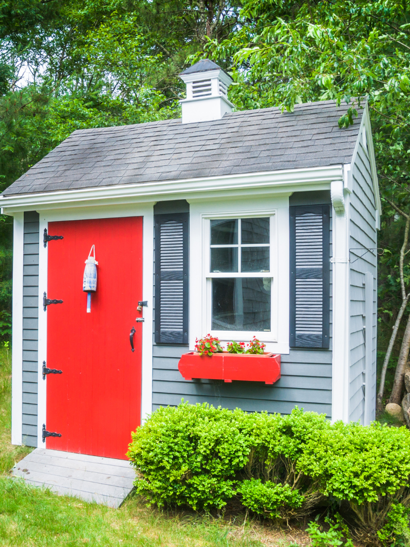 Small garden shed with red door, gray siding, and window box with flowers.