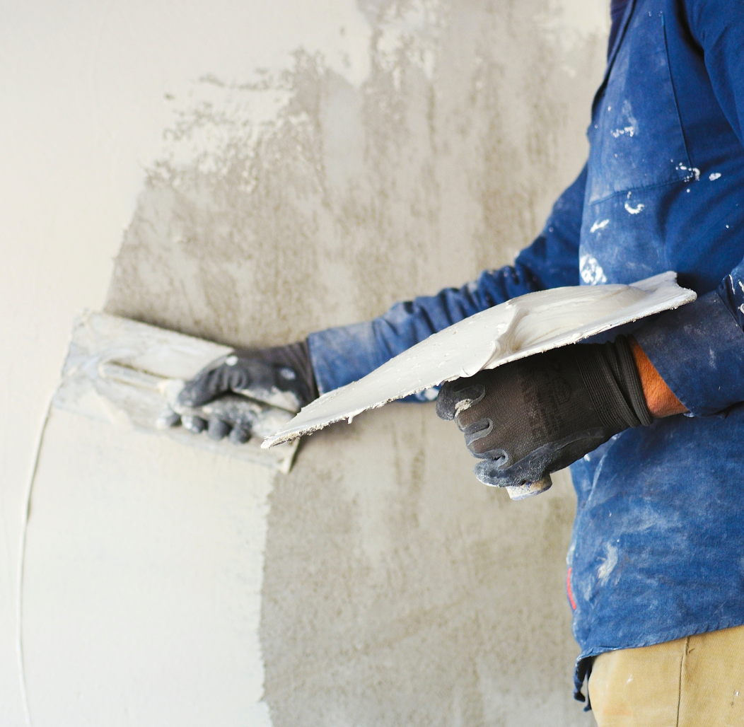 Person applying plaster to a wall with a trowel, wearing gloves and a blue shirt.