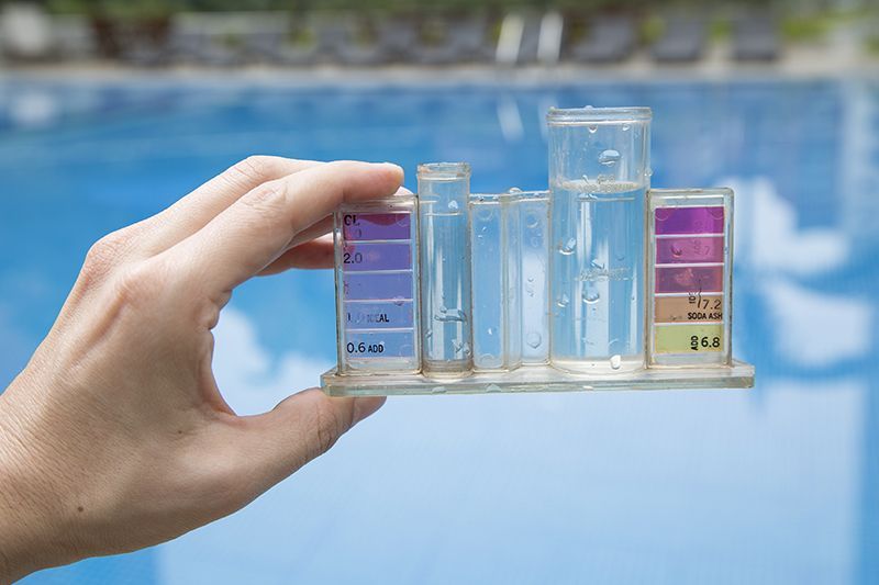 Hand holding a pool water testing kit in front of a swimming pool.