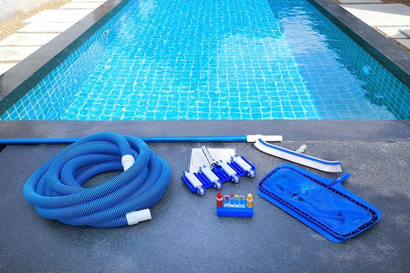 Pool cleaning equipment including vacuum hose, skimmer, and testing kit beside a swimming pool.