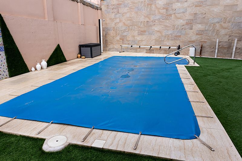 Inground swimming pool secured with a blue safety cover in a backyard.