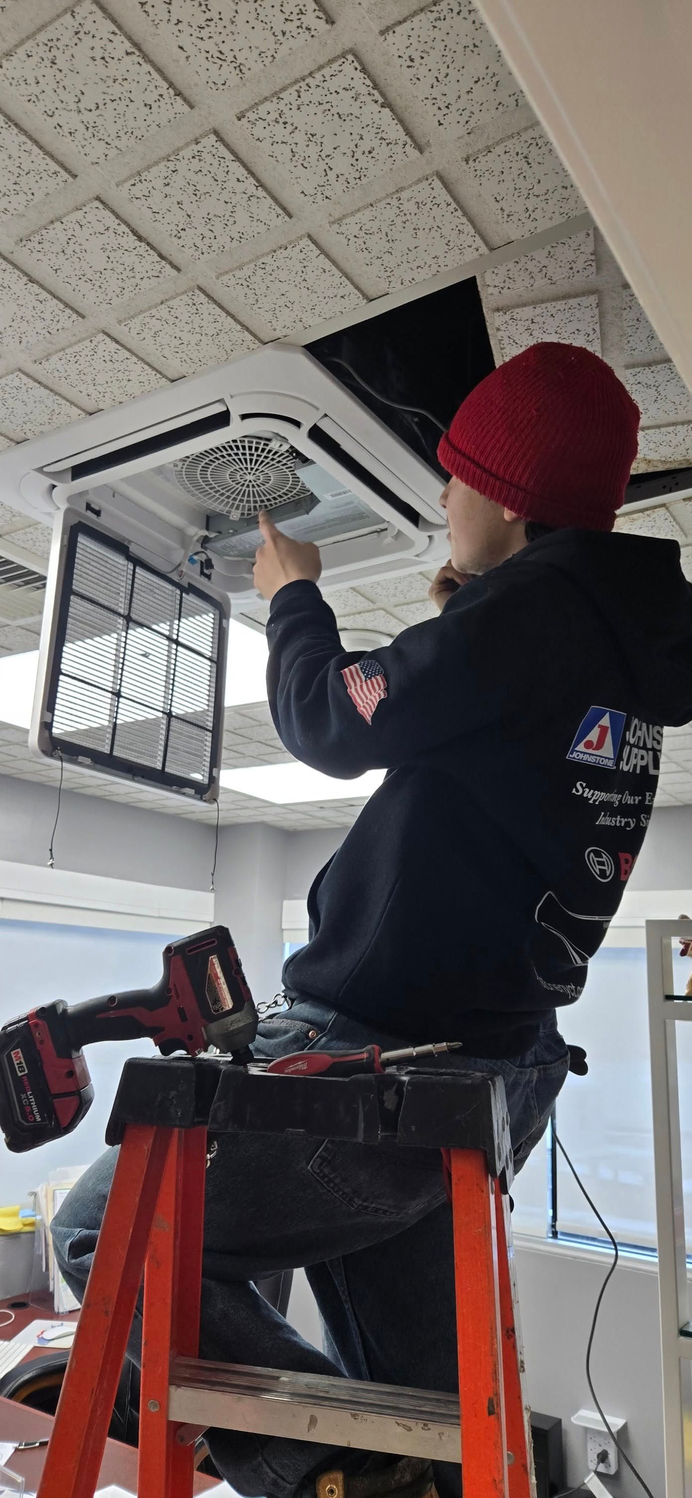 Person in red hat working on an air conditioning unit suspended from the ceiling. They are on a ladder.