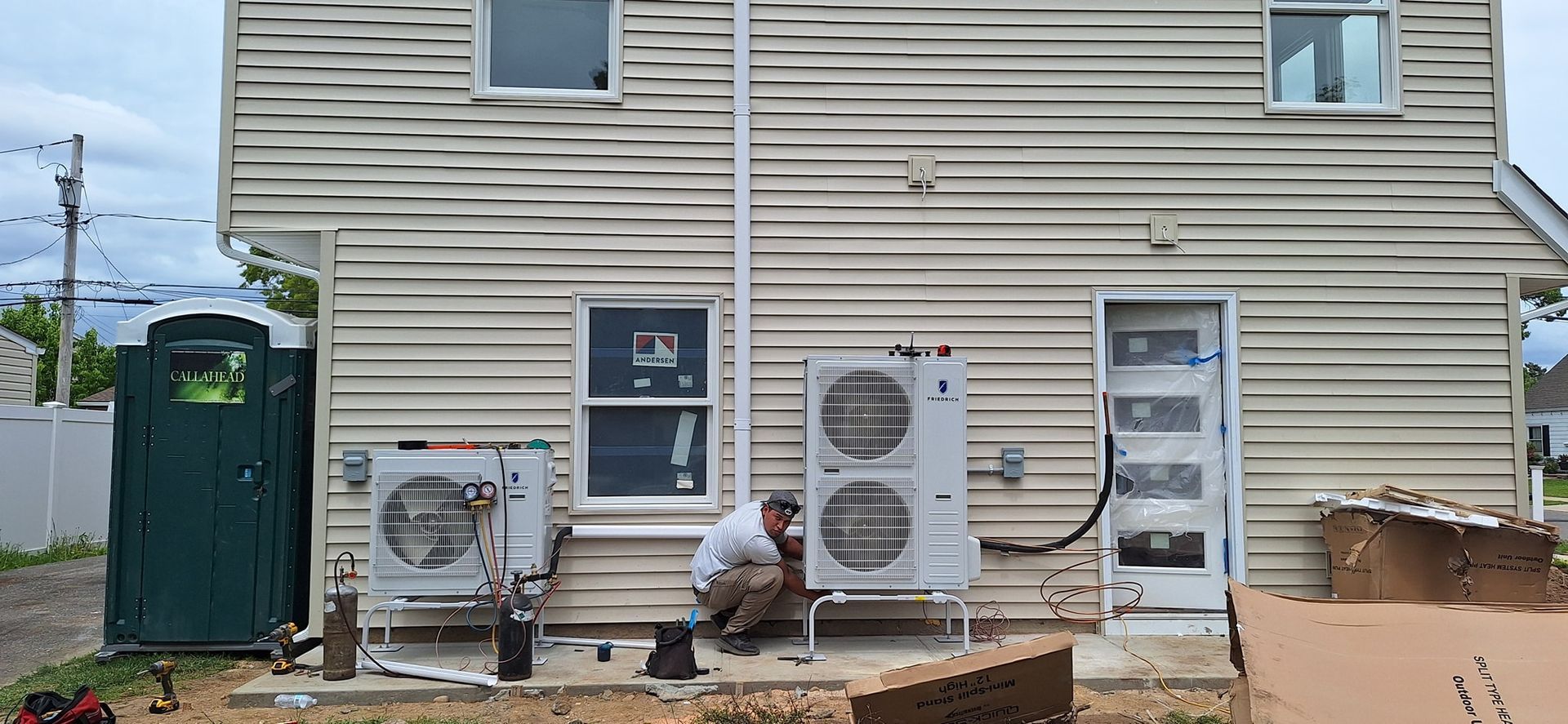 A person works on an air conditioning unit attached to a light-colored building.