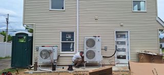 A person works on an air conditioning unit attached to a light-colored building.