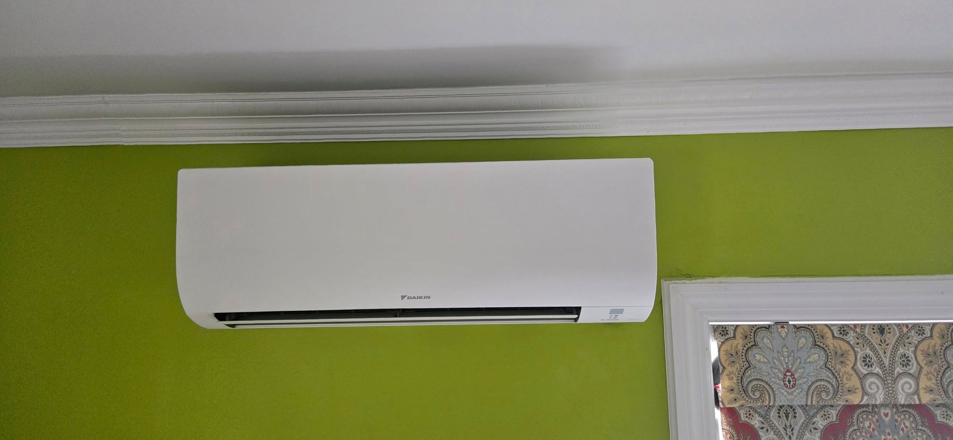 White air conditioner mounted on a green wall, near a window with a patterned curtain.