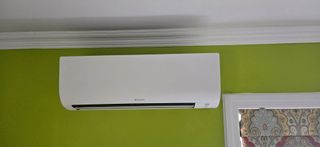White air conditioner mounted on a green wall, near a window with a patterned curtain.