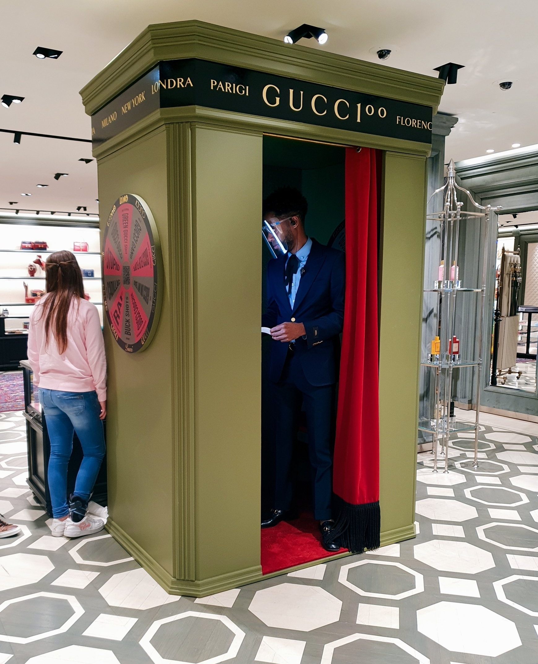 Fotohokje in een Gucci-winkel: man binnen, vrouw buiten. Olijfgroene cabine, rood gordijn, Gucci-logo.