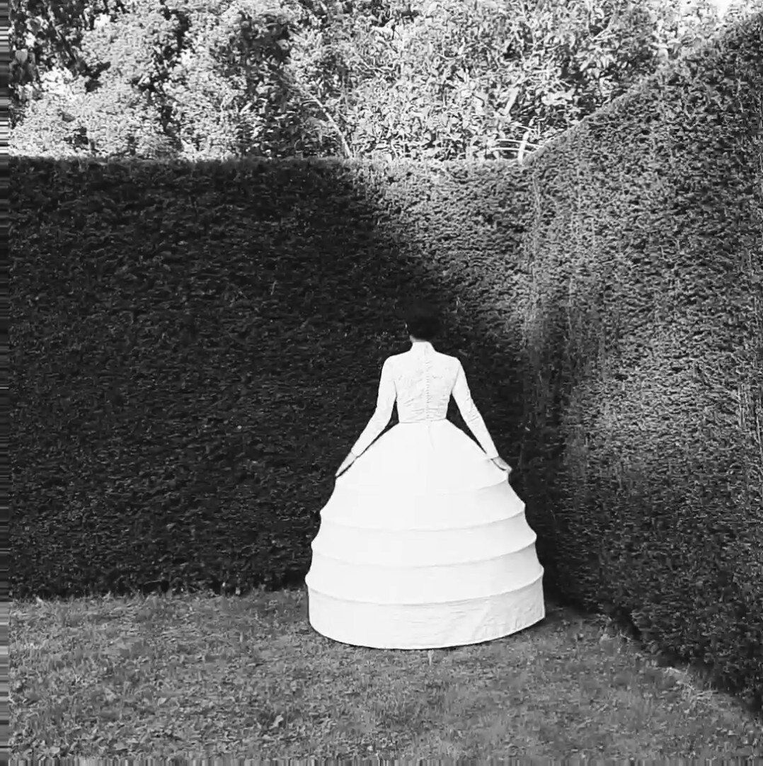 Een vrouw in een witte linnen hoepel jurk(made to order )staat in een grasveld, omringd door hoge heggen. Zwart-wit.