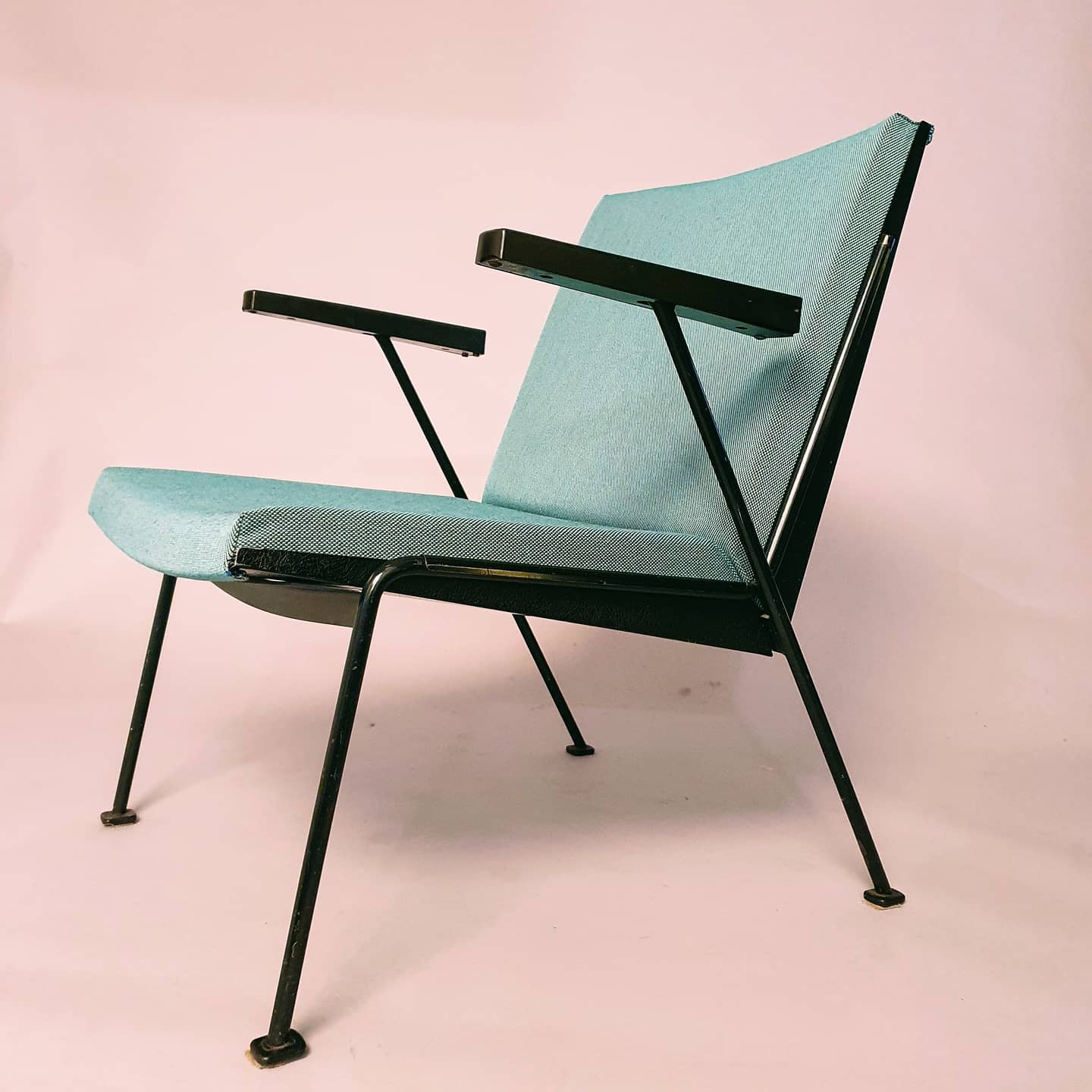 Een stoel in mid-century modern stijl met een blauwgroen kussen en een zwart metalen frame op een roze achtergrond.