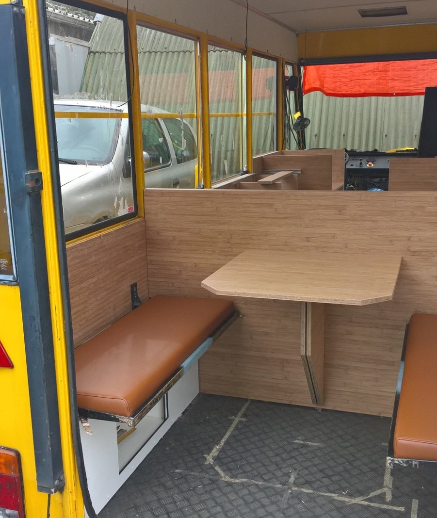 Binnenin een gele foodtruck met houten lambrisering, twee banken met bruine kussens en een uitklapbare tafel.