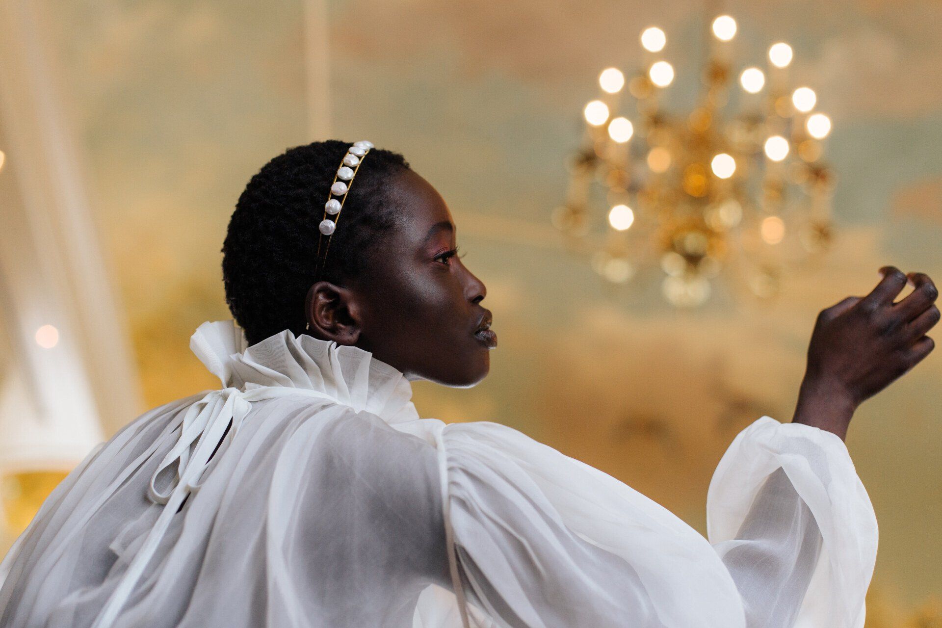 Model  met parelhaarband in een organza zijden jurk. Uitgestoken hand, omhoogkijkend, gouden kroonluchter op de achtergrond.