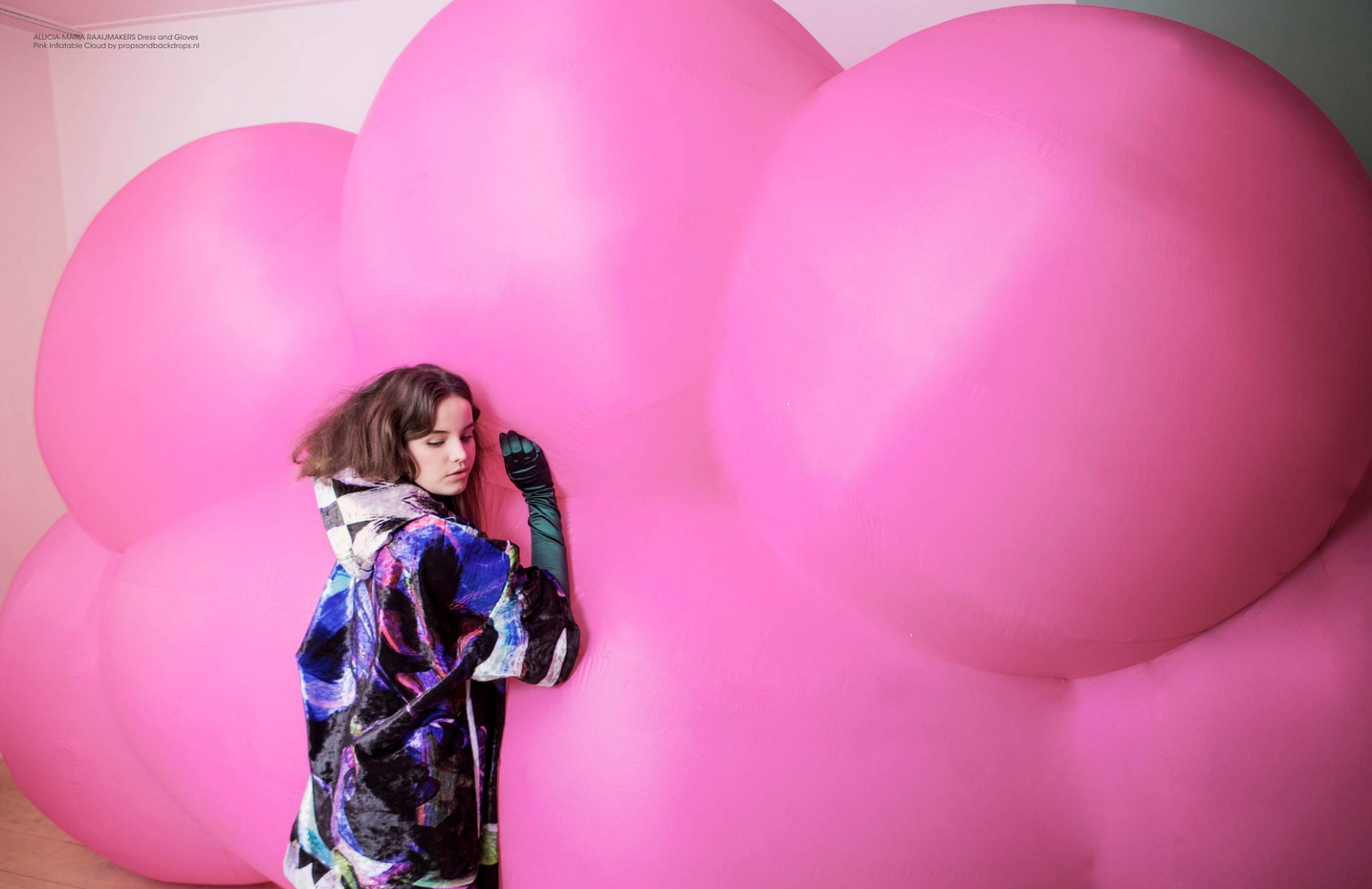 Een vrouw in een kleurrijke jas leunt tegen grote, roze, wolkachtige ballonnen inflatable  in een witte kamer.
