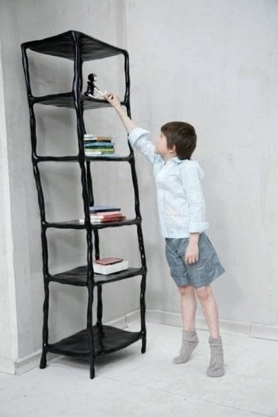 Jongen in kostuum van papier reikt naar een voorwerp van de bovenste plank in een zwarte, onregelmatige boekenkast muur.