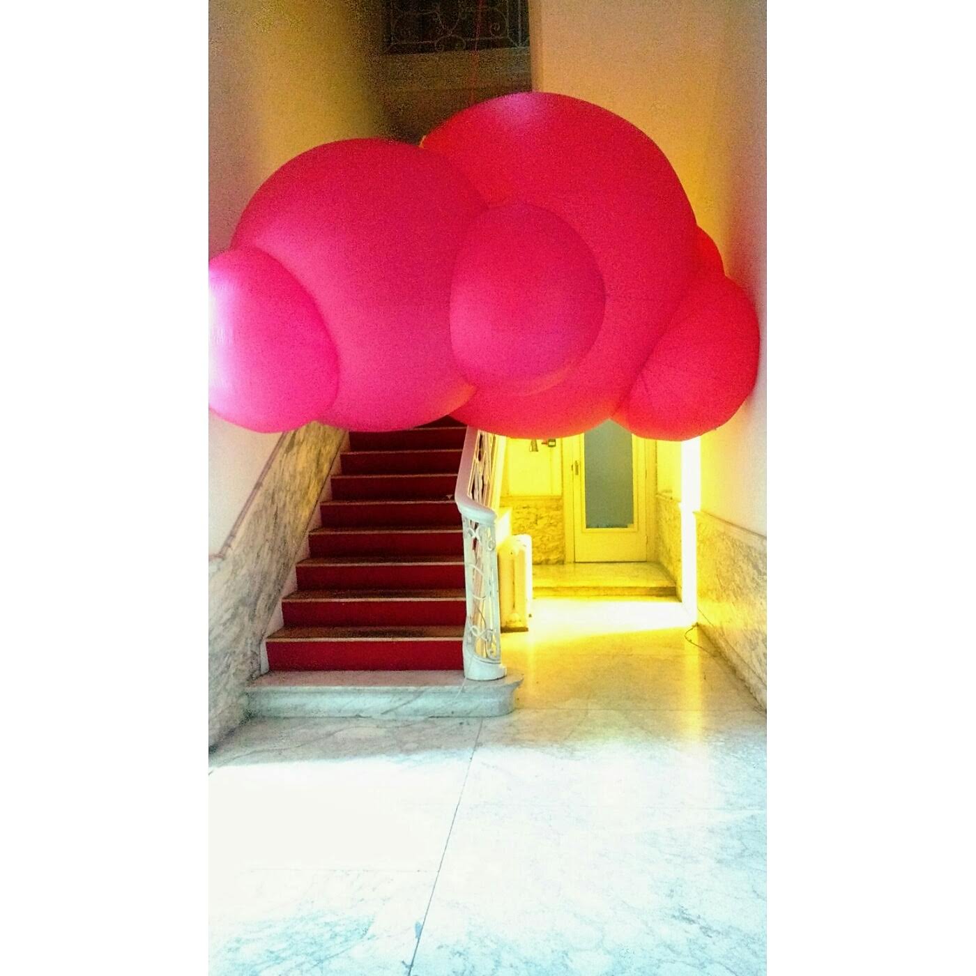 Roze, wolkachtige kunstinstallatie hangt boven een trap met rood tapijt, binnenshuis.