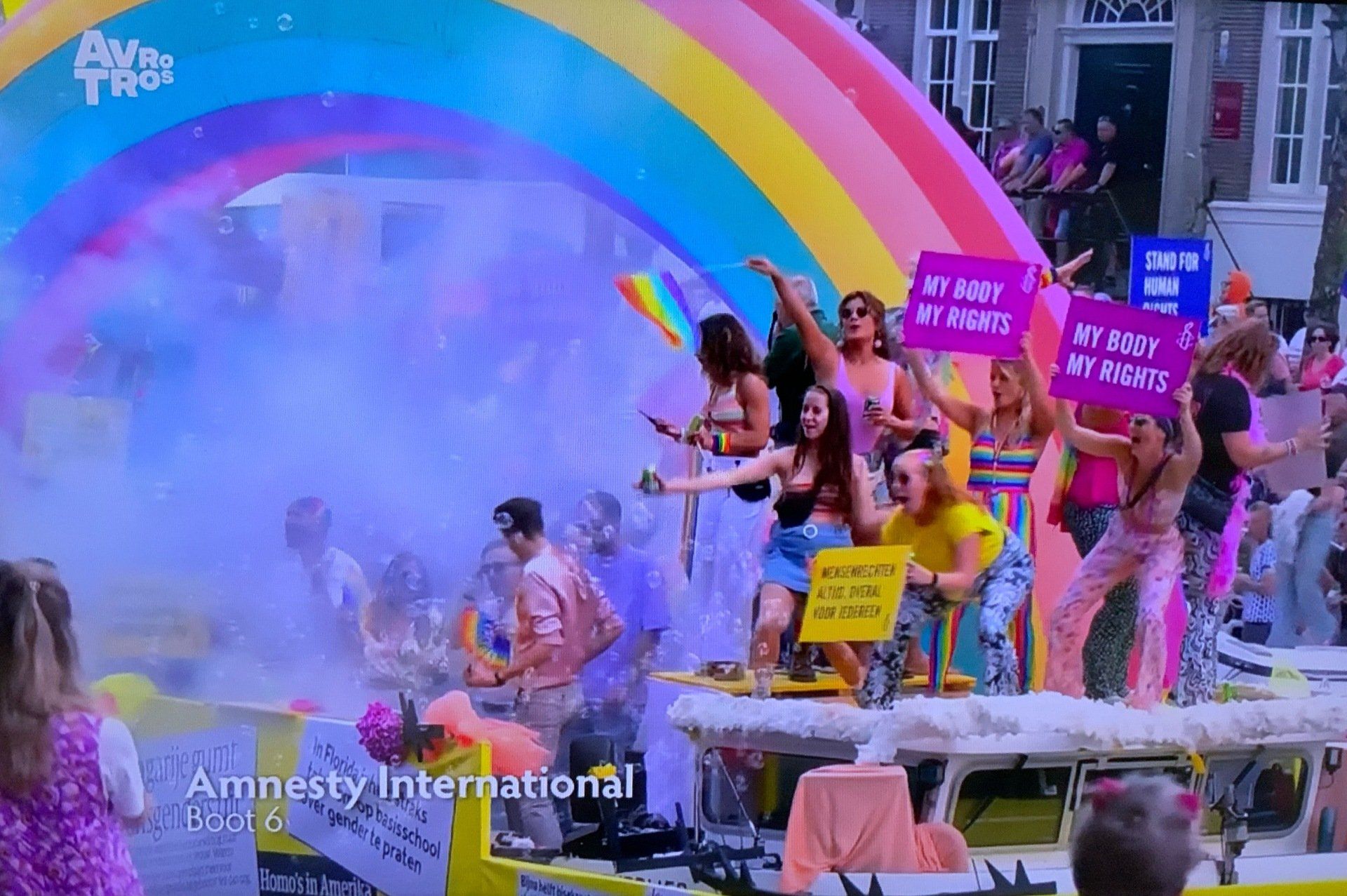 Mensen vieren Pride op een praalwagen met een regenboog. Ze houden protest borden vast. 