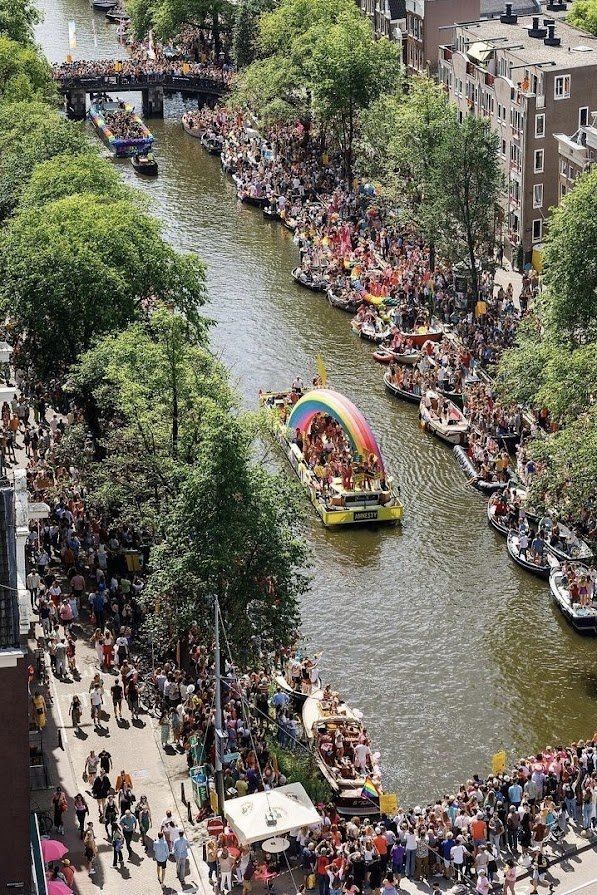 Amsterdamse gracht tijdens Pride-parade met kleurrijke praalwagens, drukte en regenboogvlag.