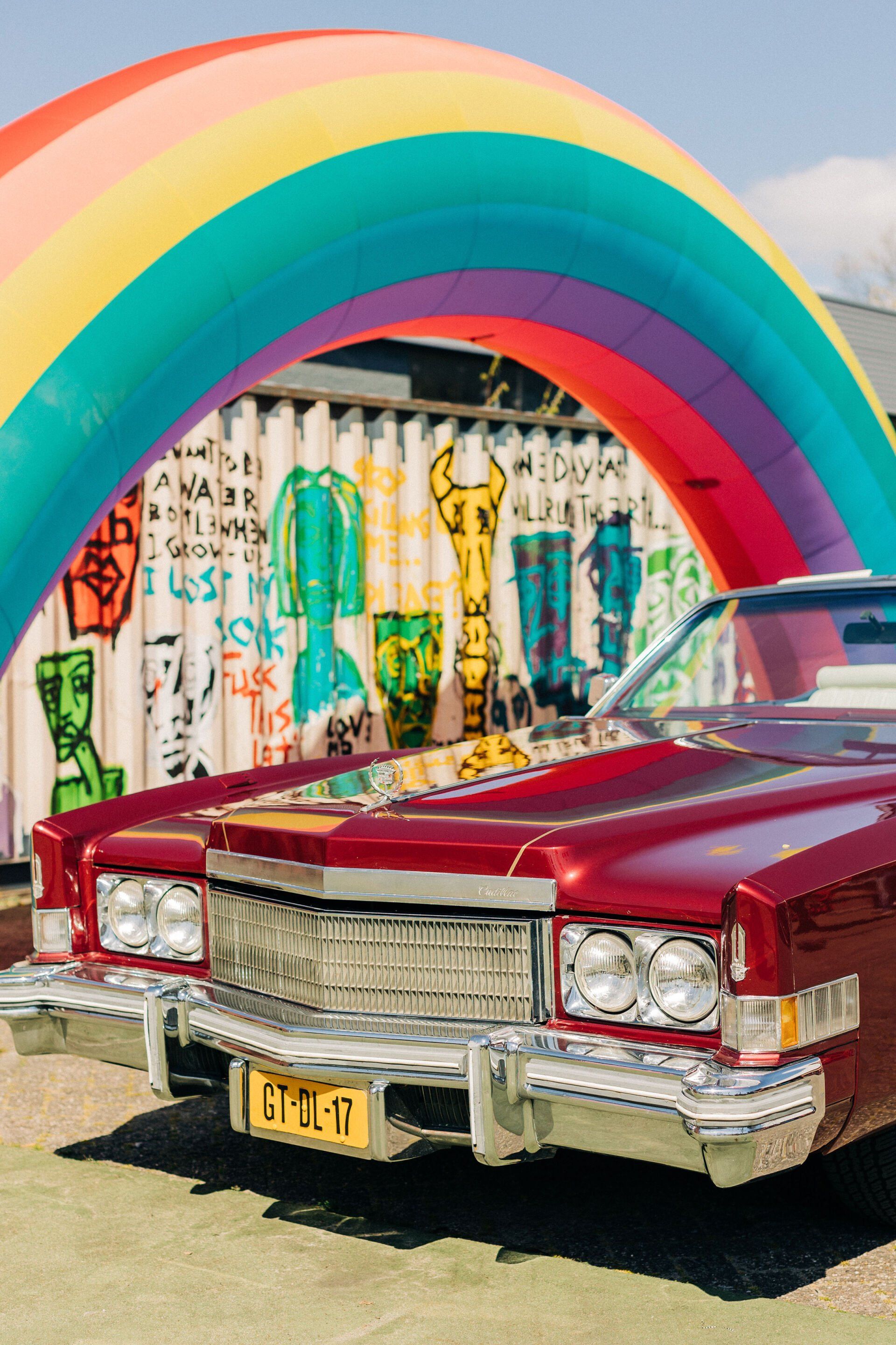 Een klassieke rode auto geparkeerd onder een regenboogboog, voor een met graffiti bedekt gebouw / muur.