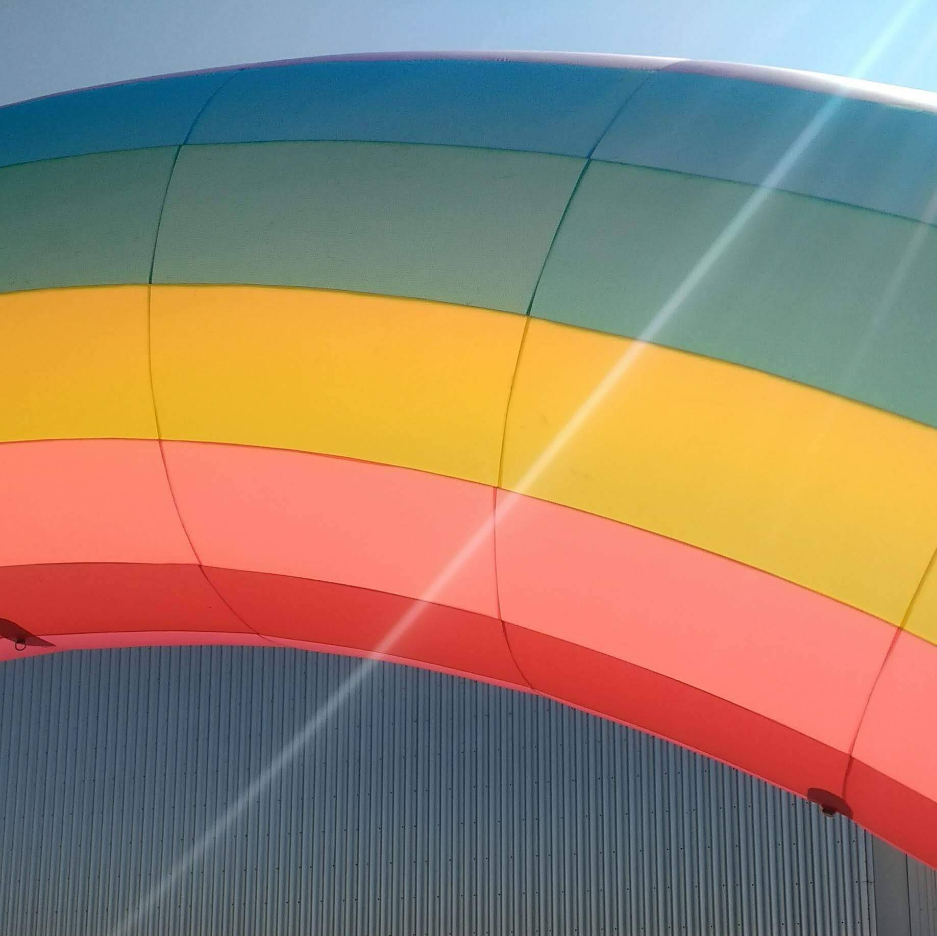 Een luchtballon met regenboogstrepen tegen een blauwe lucht en een metalen gebouw met structuur.