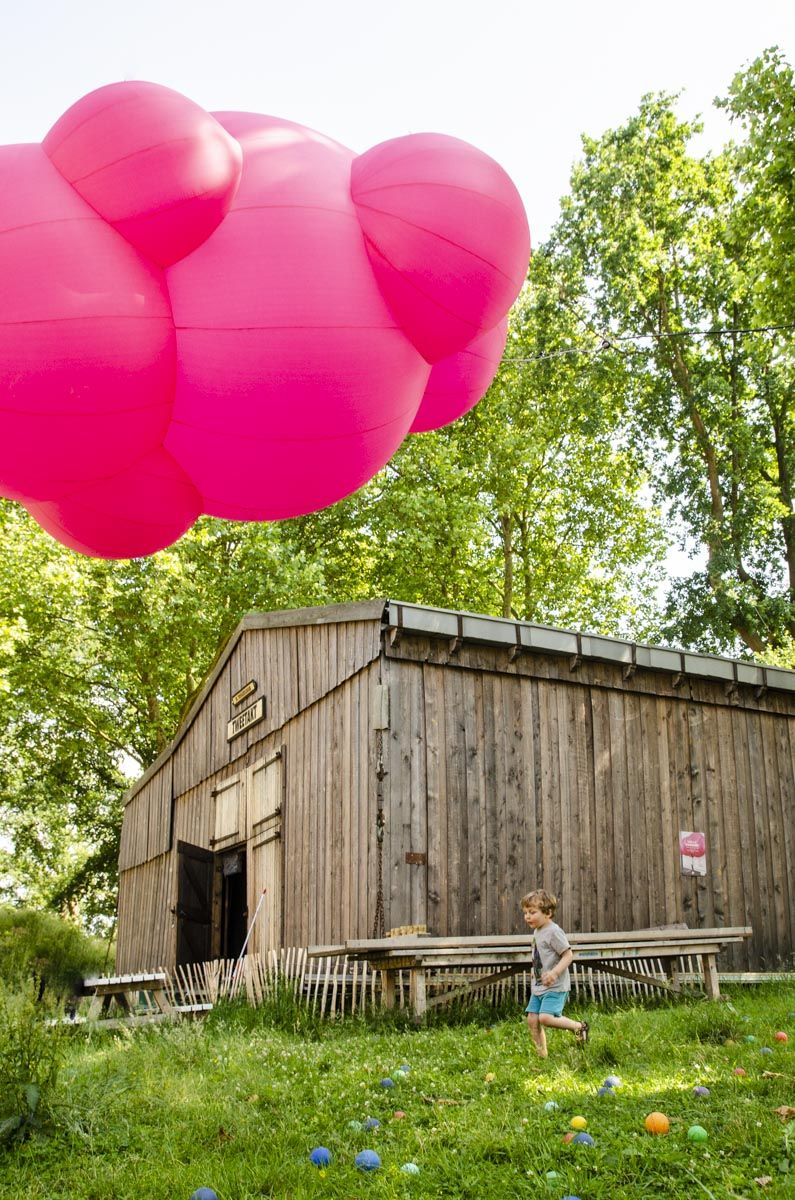 Een felroze ballon zweeft boven een houten hut terwijl een kind door een grasveld rent.