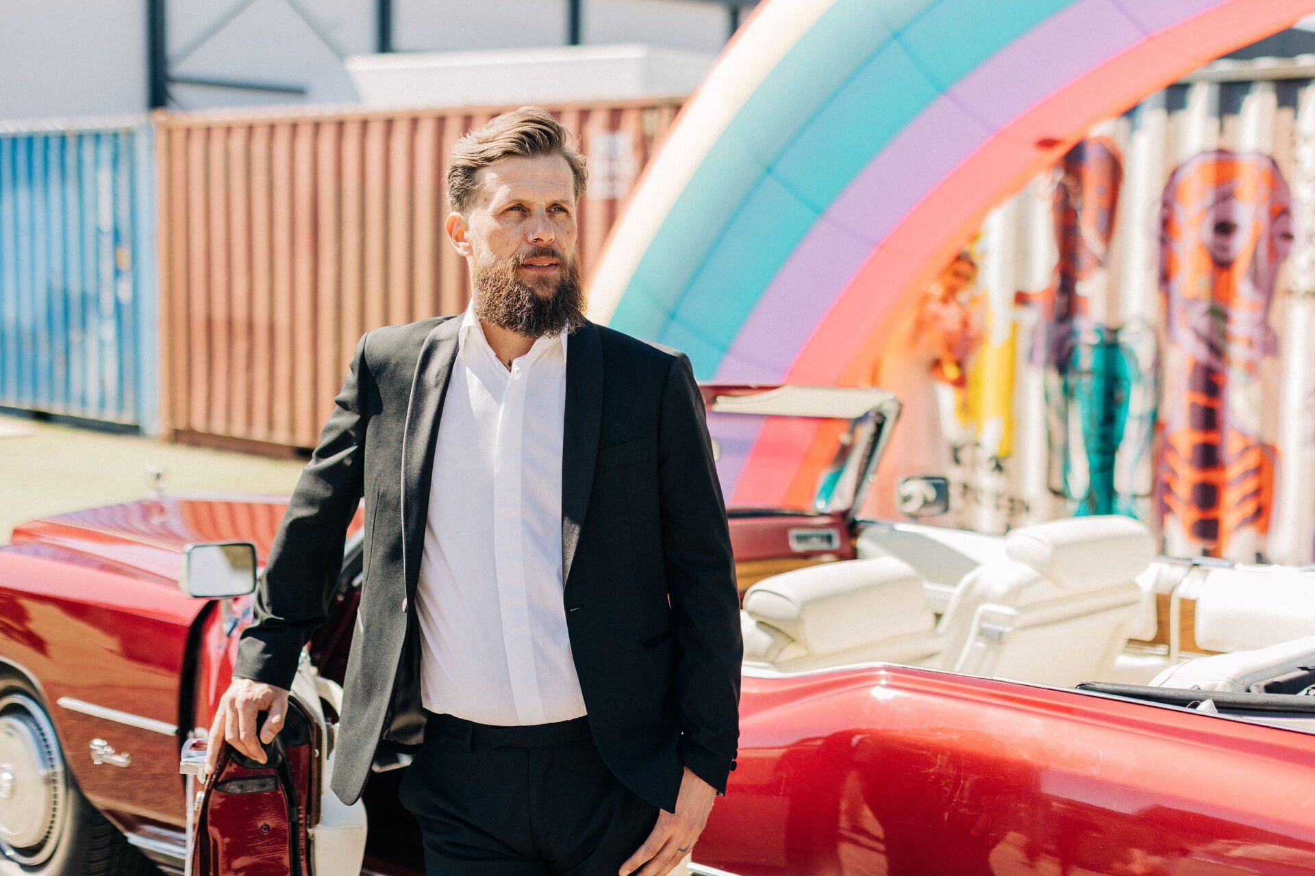 Een man in een zwarte blazer staat naast een rode cabriolet onder een kleurrijke regenboogboog.