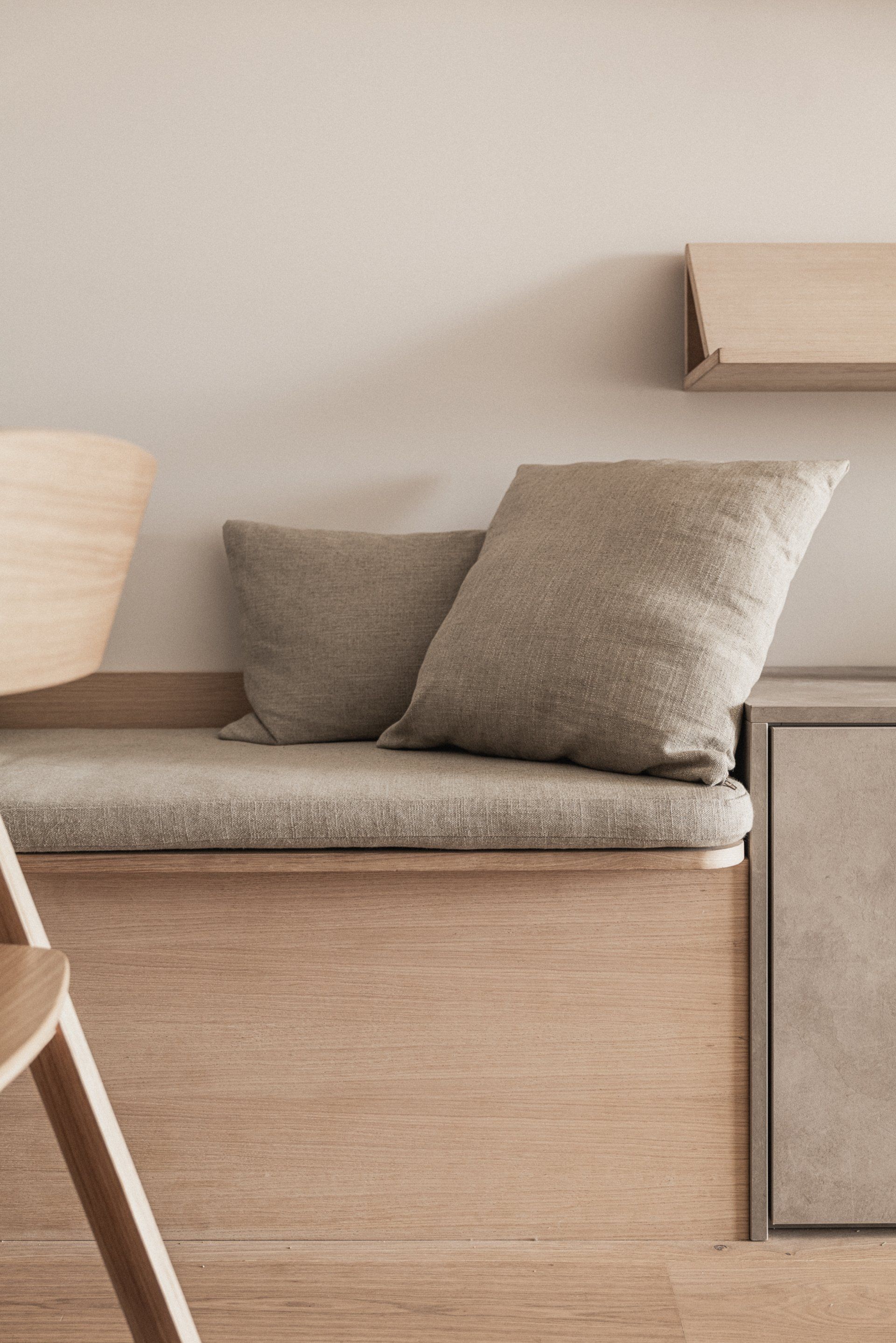 Houten  gestoffeerde bank met twee kussens tegen een beige muur, naast een lichtgekleurde kast.