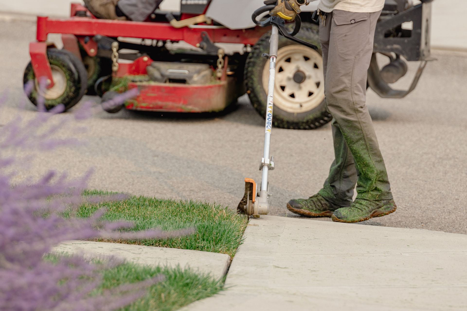 A person is standing on a sidewalk cutting grass with a lawn mower in the background.