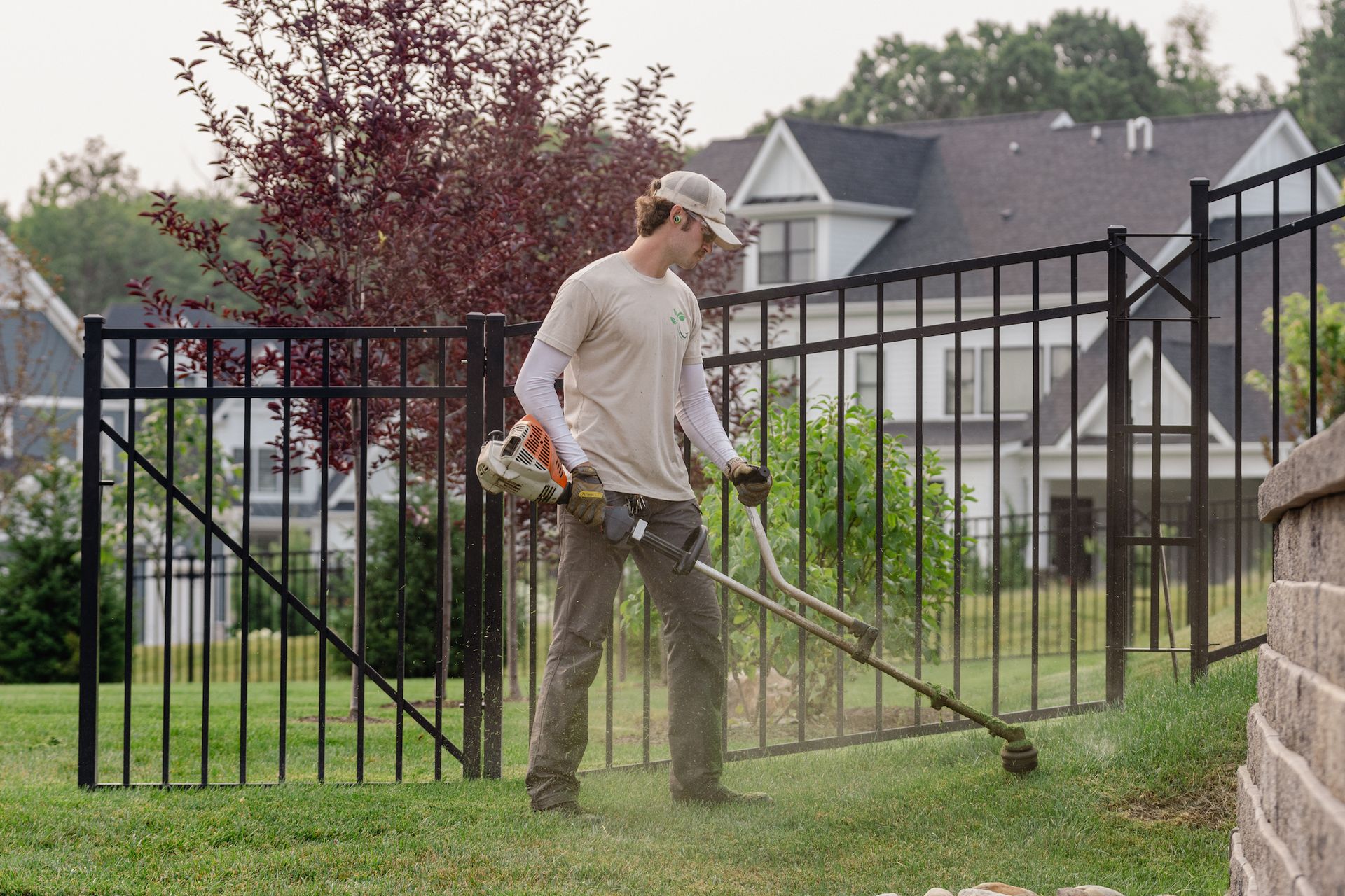 A man is using a lawn mower to cut the grass in front of a fence.