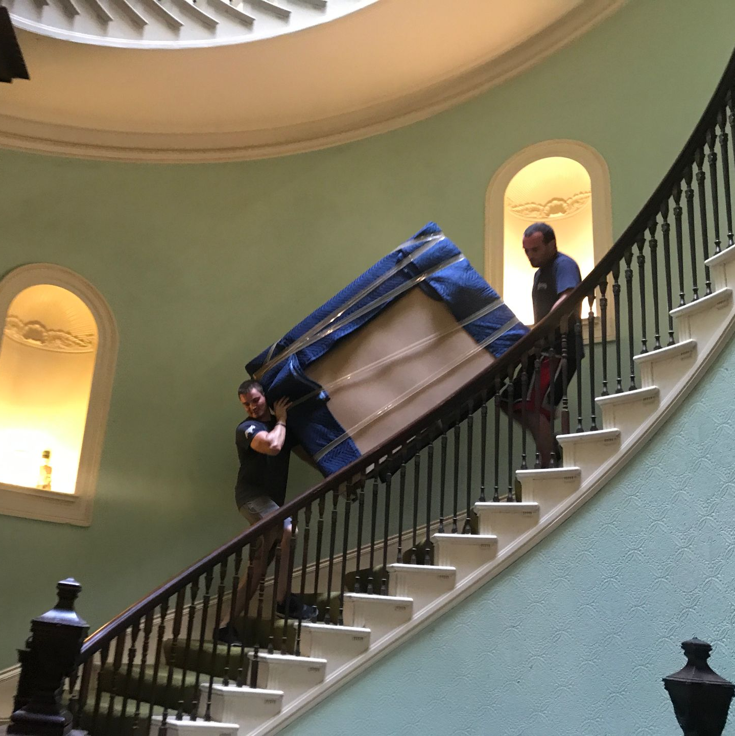 Two people carrying a piano up a curved staircase in a building. The piano is wrapped in a blue blanket.