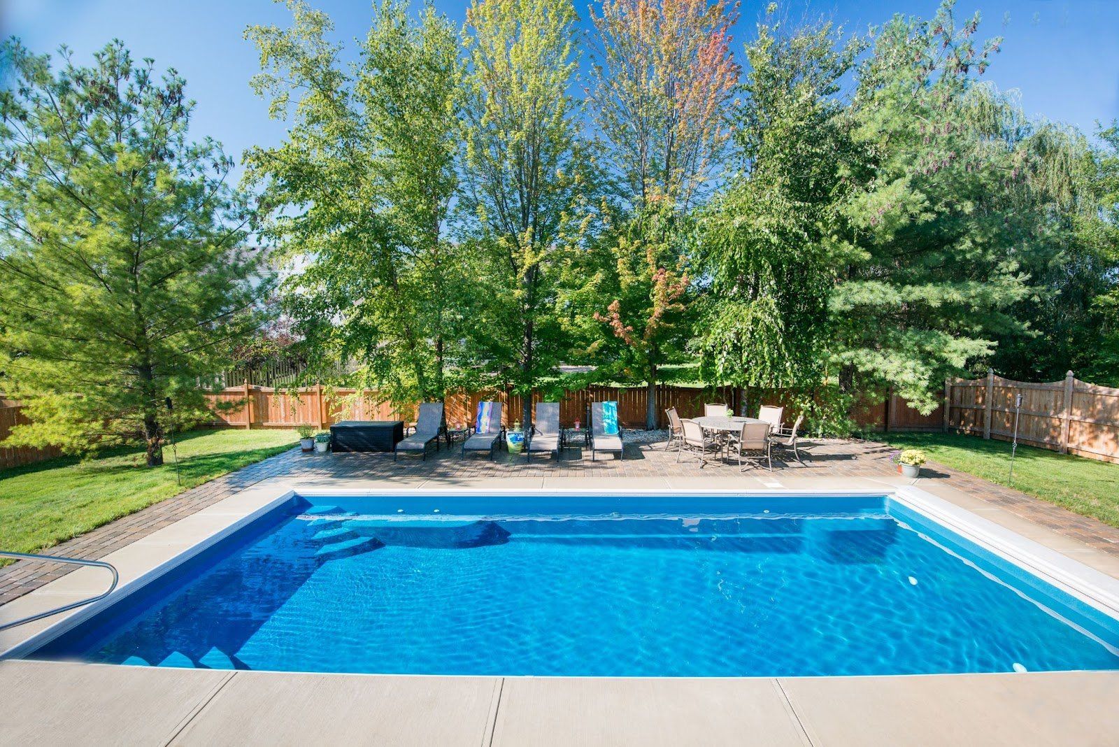 Swimming pool with lounge chairs, table, and trees in a backyard on a sunny day.