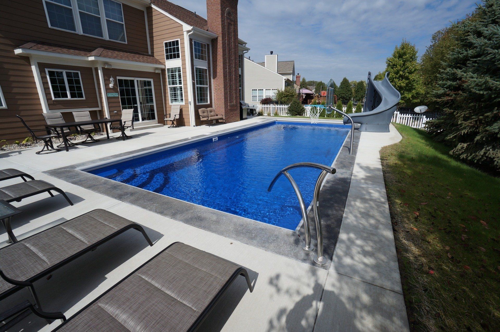 Rectangular blue swimming pool next to a house with lounge chairs on the concrete patio.