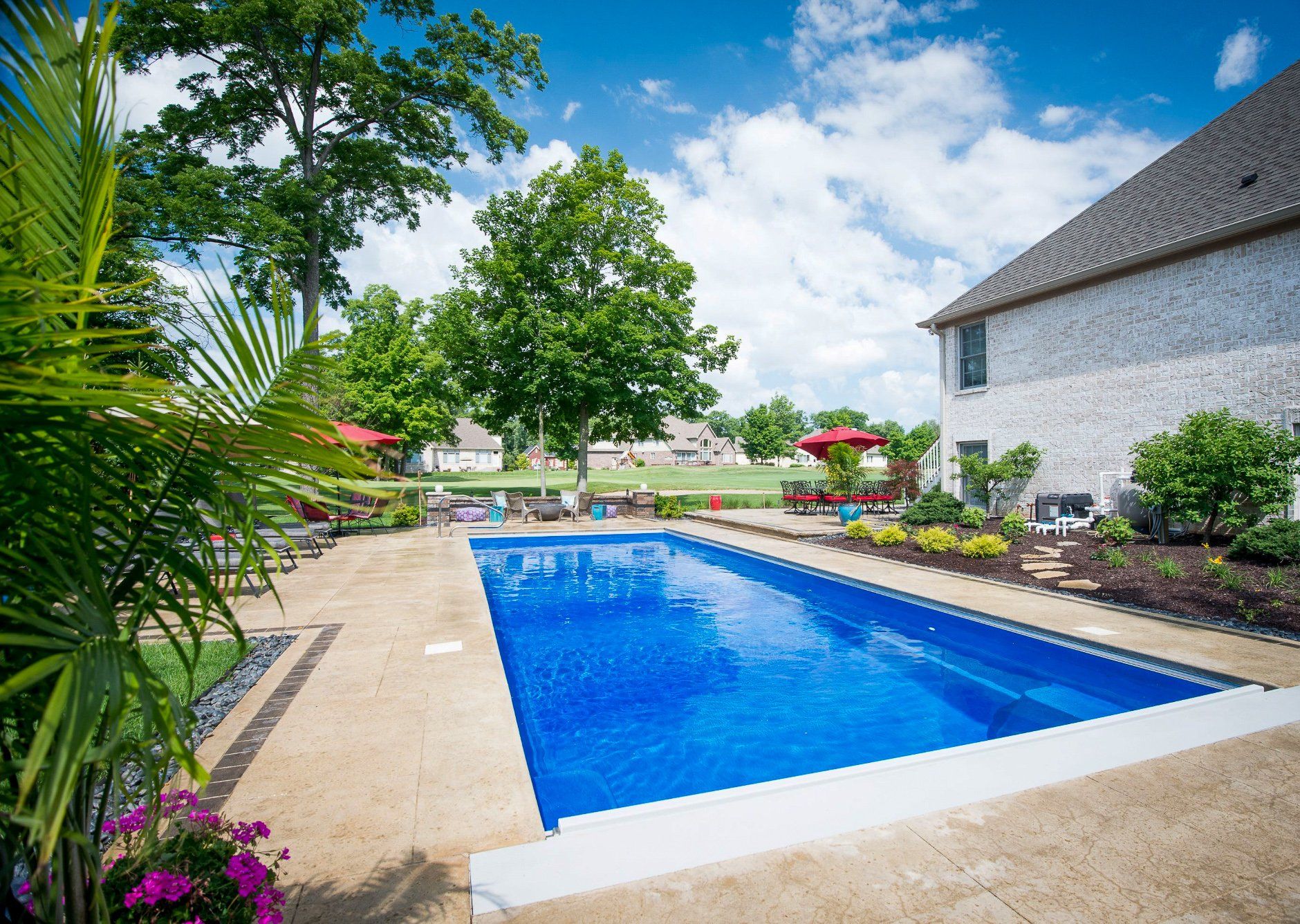 Rectangular pool in backyard, blue water, beige patio, house with brick siding, sunny day.