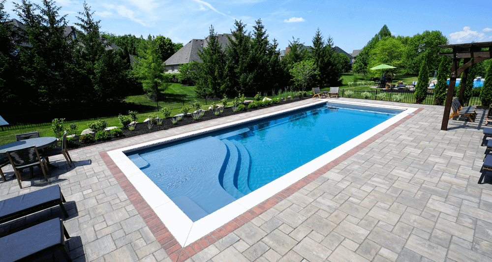 Rectangular blue pool with steps, surrounded by stone patio, green yard, and houses under a blue sky.